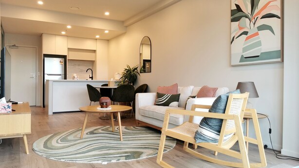 Ein modernes Wohnzimmer mit einer Couch, einem Couchtisch und einem Schaukelstuhl. Im Hintergrund ist eine Küchenzeile mit einem Kühlschrank und einer Spüle zu sehen.