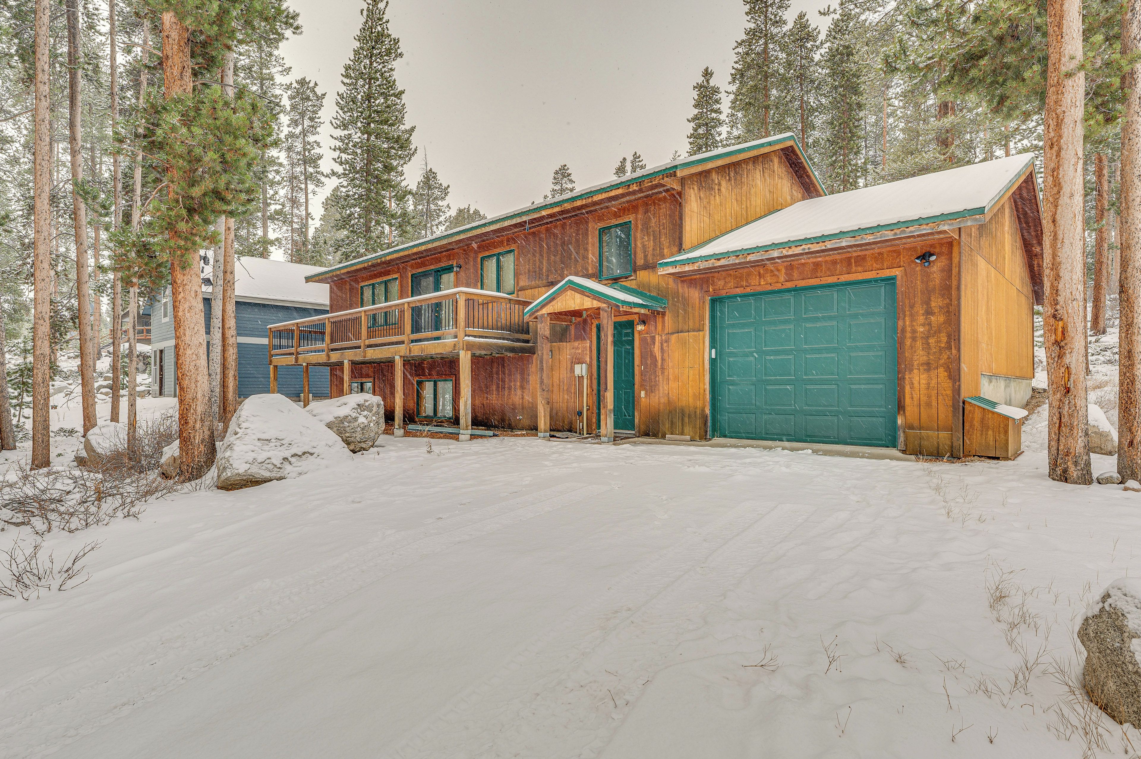 A wooden cabin with a green garage door, surrounded by snow-covered ground and tall pine trees.