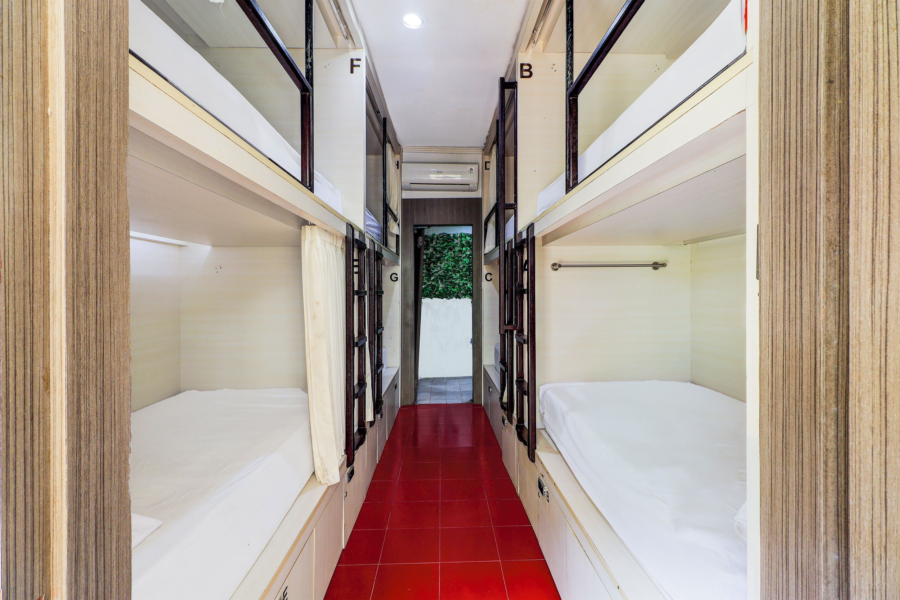A narrow corridor with bunk beds on either side, a red-tiled floor, and a door at the end leading to a green outdoor area.