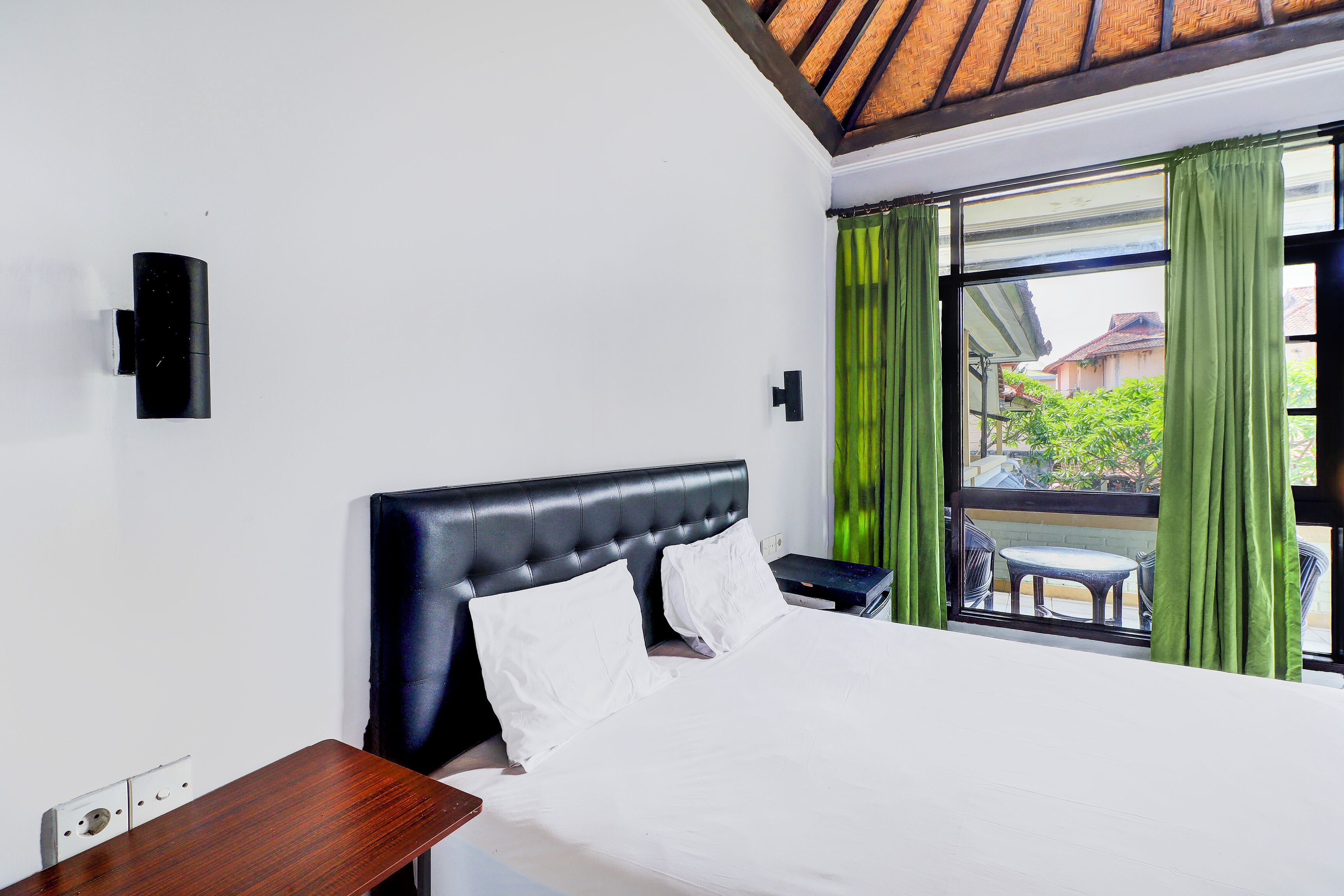 A hotel room with a bed, a nightstand, a wall-mounted lamp, and a window with green curtains.