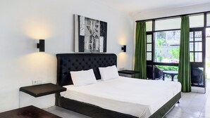 A hotel room with a large bed, a wall-mounted artwork, a small table, and a view through a window with curtains. - Collection O Legian Near Bimc Hospital Nusa Uda Formerly Anna Kuta Inn (Kuta)