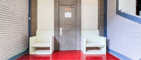 Un pasillo con suelo de baldosas rojas, una puerta de madera con el número 7, y dos bancos blancos a cada lado.