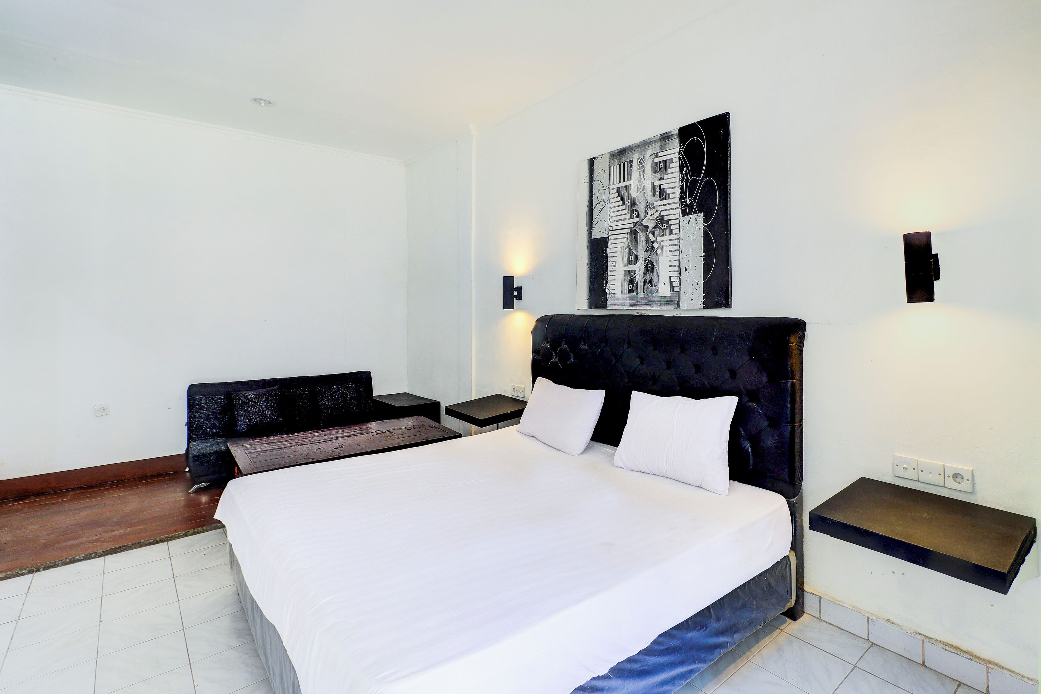 A hotel room with a large bed, a headboard, a wall-mounted artwork, a bedside table, and a bench.