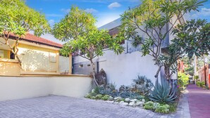 A courtyard with a paved walkway, a building with a red-tiled roof, and various plants and trees. - Collection O Legian Near Bimc Hospital Nusa Uda Formerly Anna Kuta Inn (Kuta)