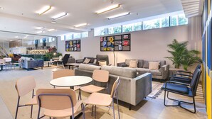 Un espace de détente moderne, doté de canapés, de fauteuils et de tables. De grandes fenêtres laissent entrer la lumière naturelle, et un mur est décoré de divers panneaux et tableaux.