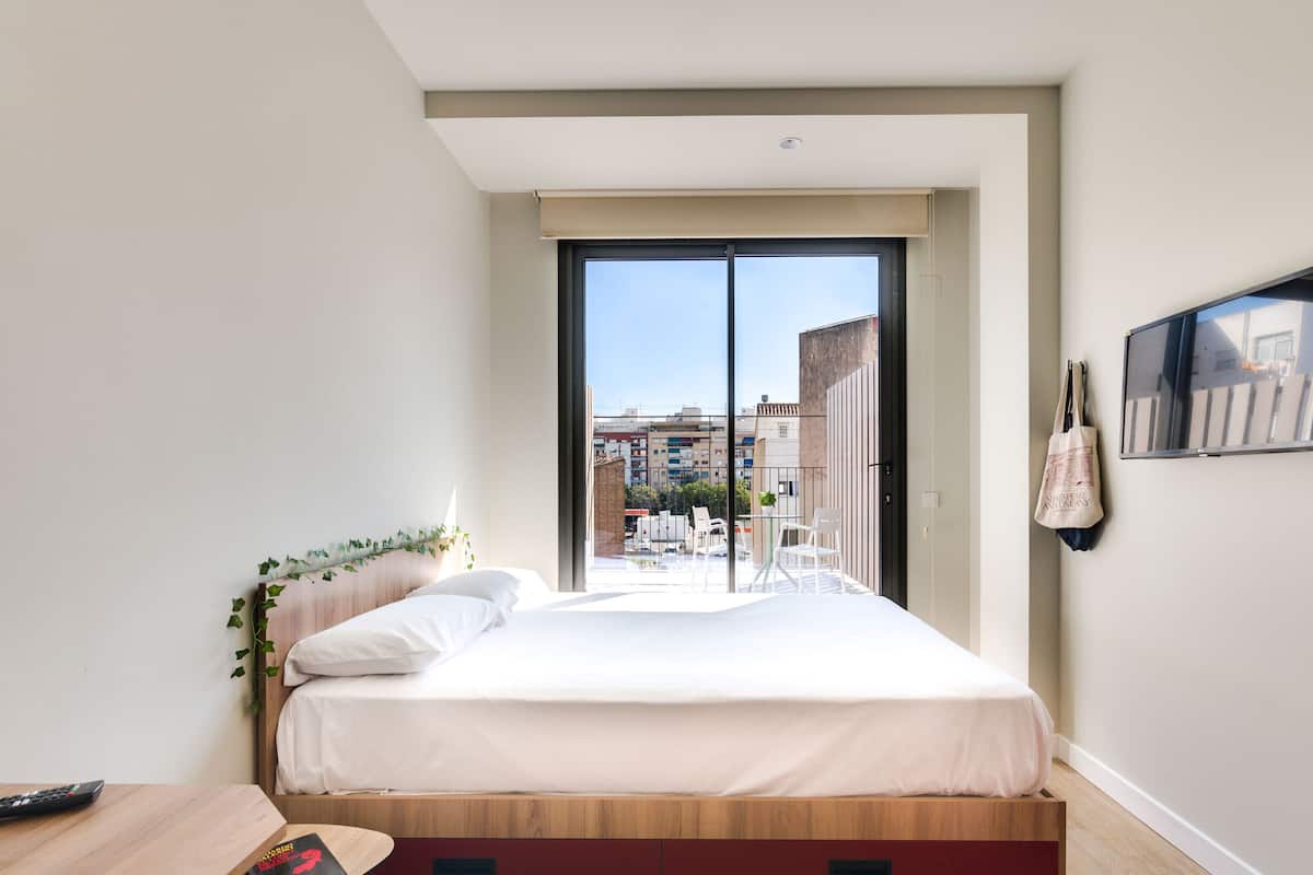 A modern bedroom with a large bed, a wooden desk, a TV, and a balcony with a city view.