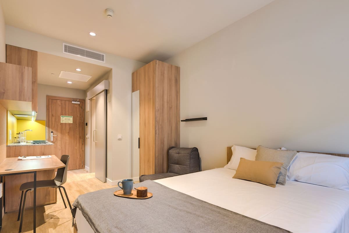 A modern hotel room with a bed, a desk with a chair, a small table with a cup, and a chair.