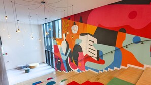 Un intérieur moderne agrémenté d’une fresque murale abstraite aux couleurs vives, d’un parquet et de coussins aux motifs géométriques, ainsi que de luminaires suspendus.