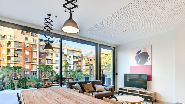 Un salon moderne avec une table à manger en bois, un canapé et un téléviseur. De grandes fenêtres offrent une vue sur un immeuble résidentiel et sur la verdure.