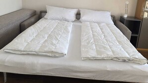 Una cama doble con ropa de cama blanca, un sofá gris, una mesita de noche y un buró.