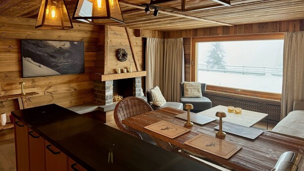 L’intérieur chaleureux d’un chalet avec un plafond en bois, une cheminée, un coin repas avec une table et des chaises, et une grande fenêtre donnant sur un paysage enneigé.