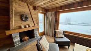 L’intérieur chaleureux d’un chalet avec une cheminée en pierre, des murs en bois et une grande fenêtre donnant sur un paysage enneigé.