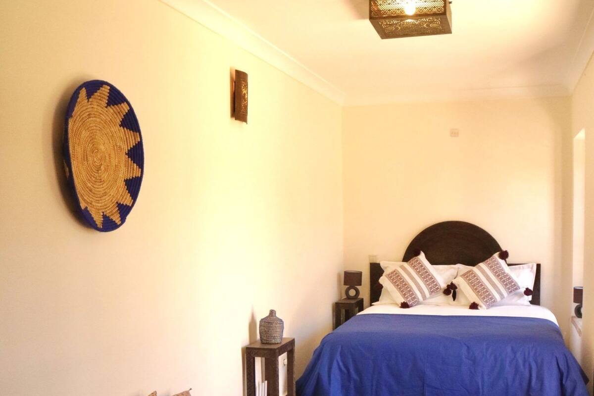 A room with two beds, each with a blue bedspread and pillows, a small table with a vase, and a wall hanging.