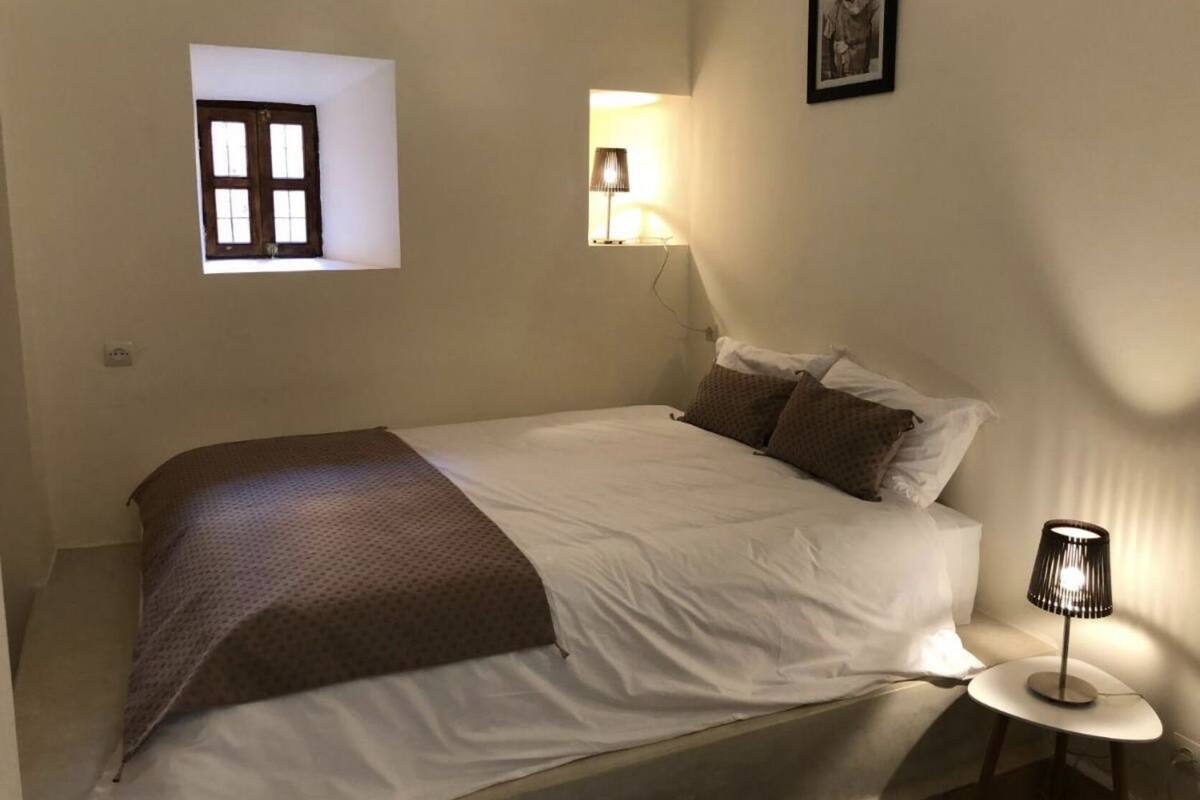 A bedroom with a bed, a small window, a wall-mounted lamp, and a framed picture.
