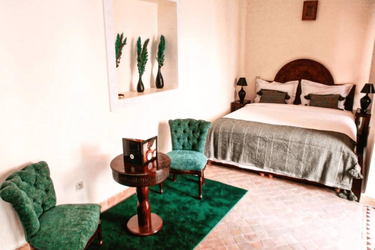 A bedroom with a bed, two green armchairs, a small round table, and a green rug.