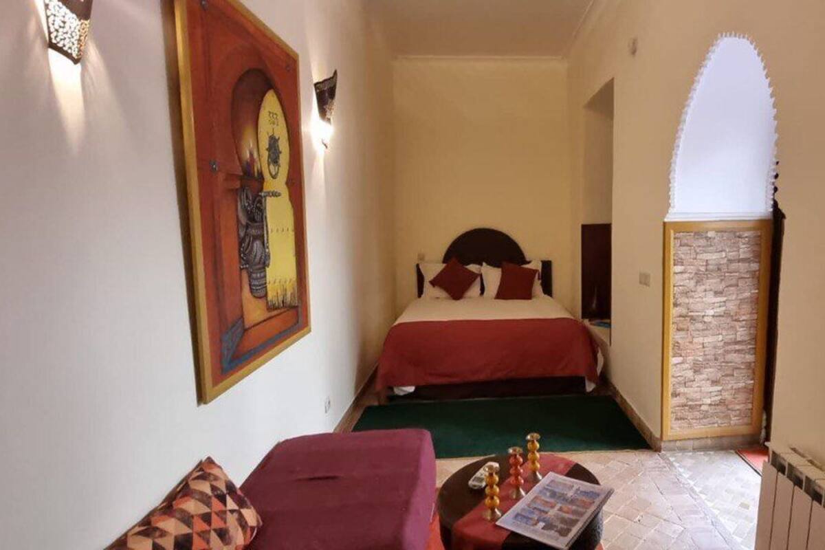 A narrow hallway leading to a bed with a red bedspread, a small table with a magazine, and a wall-mounted light fixture.