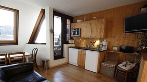 Een compacte keuken met houten kastjes, een magnetron en een koffiezetapparaat. In de woonkamer staat een leren zwarte bank en een rieten fauteuil.