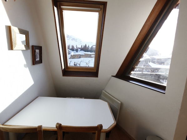 Une pièce d’angle avec une table blanche, un radiateur et une fenêtre donnant sur un paysage enneigé.