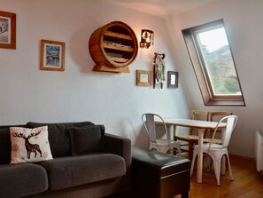 A cozy living room with a grey sofa, a wooden barrel wall decoration, a dining table with chairs, and a large window offering a view.