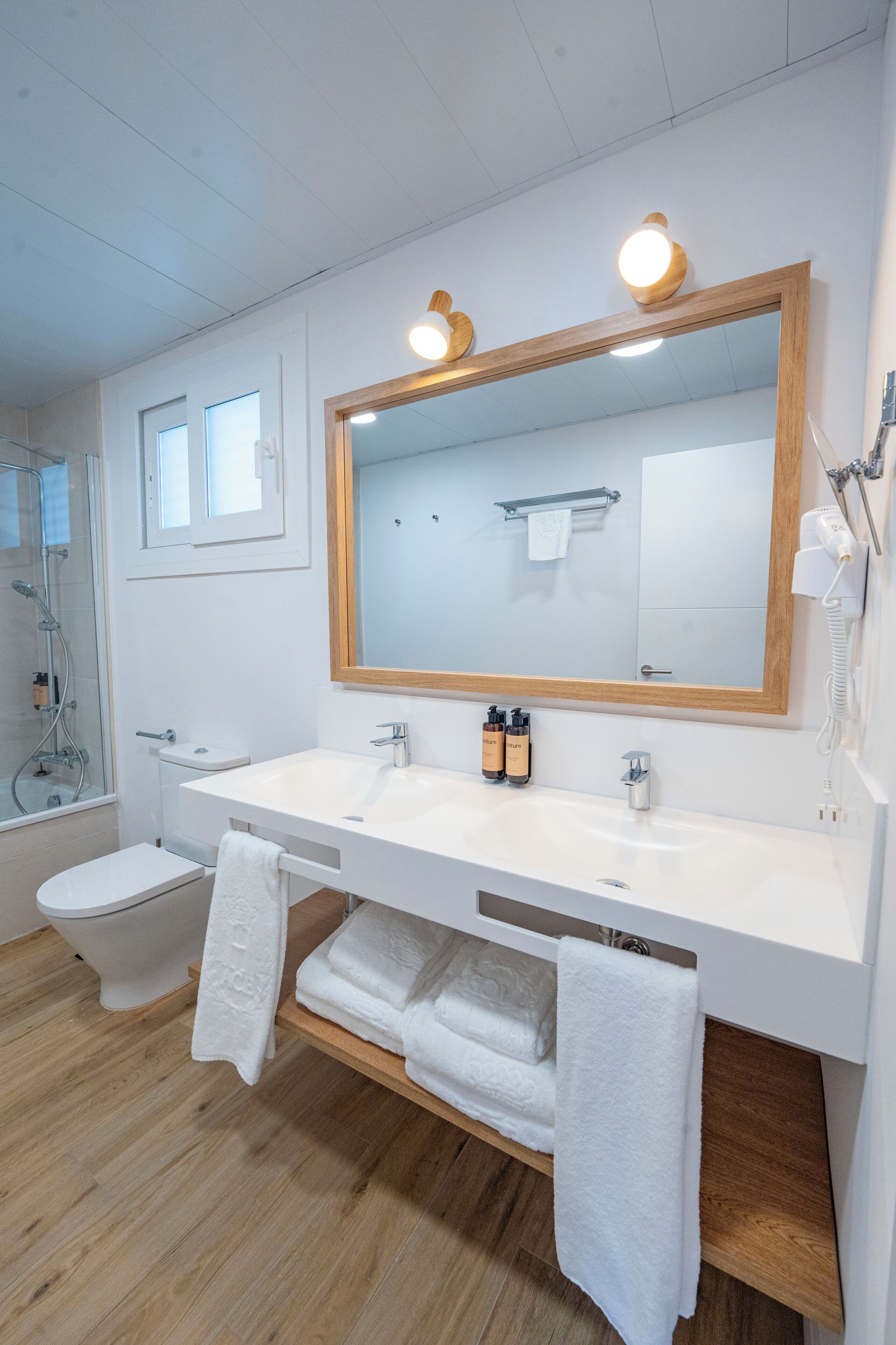 A modern bathroom with a large mirror, white sink, and toilet. There are towels on the floor and a hairdryer on the wall.