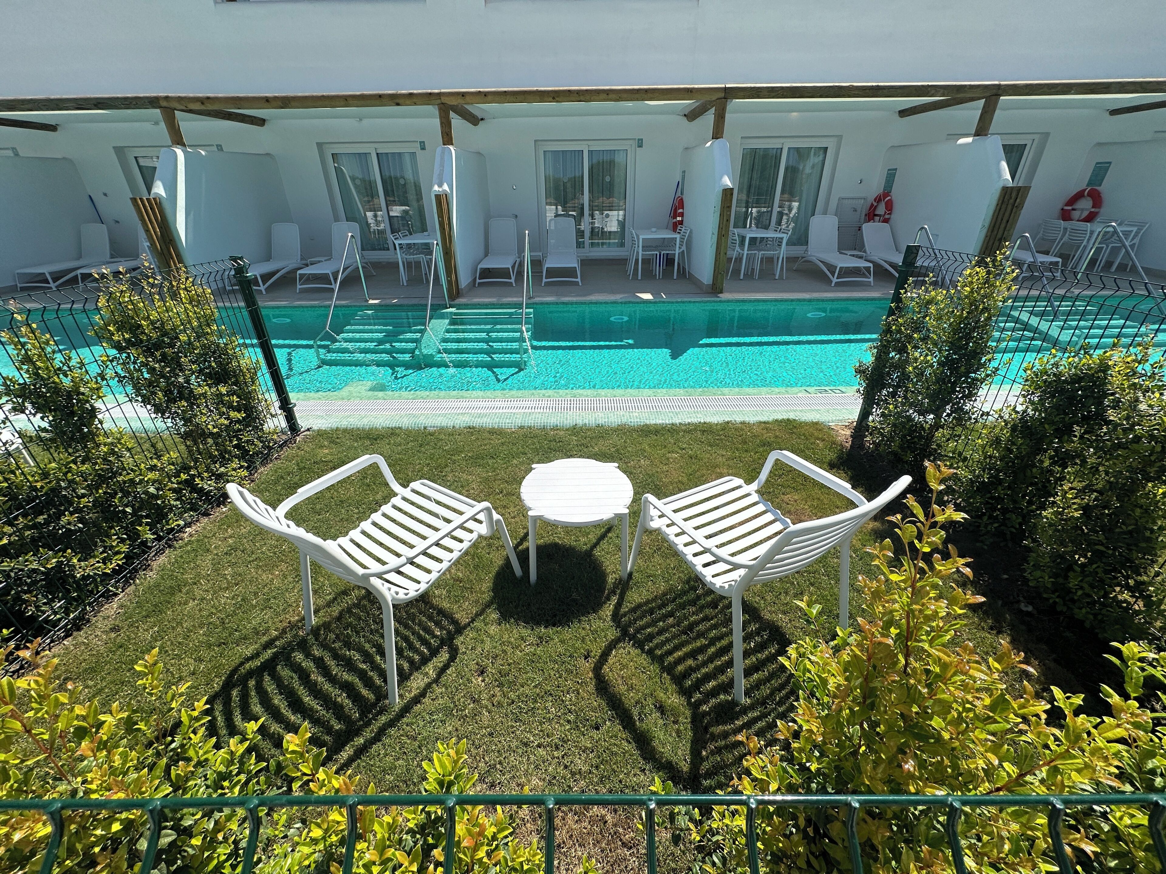 Un espace aménagé au bord de la piscine, avec des chaises longues blanches et une petite table, le tout entouré d’une pelouse et d’arbustes bien entretenus.