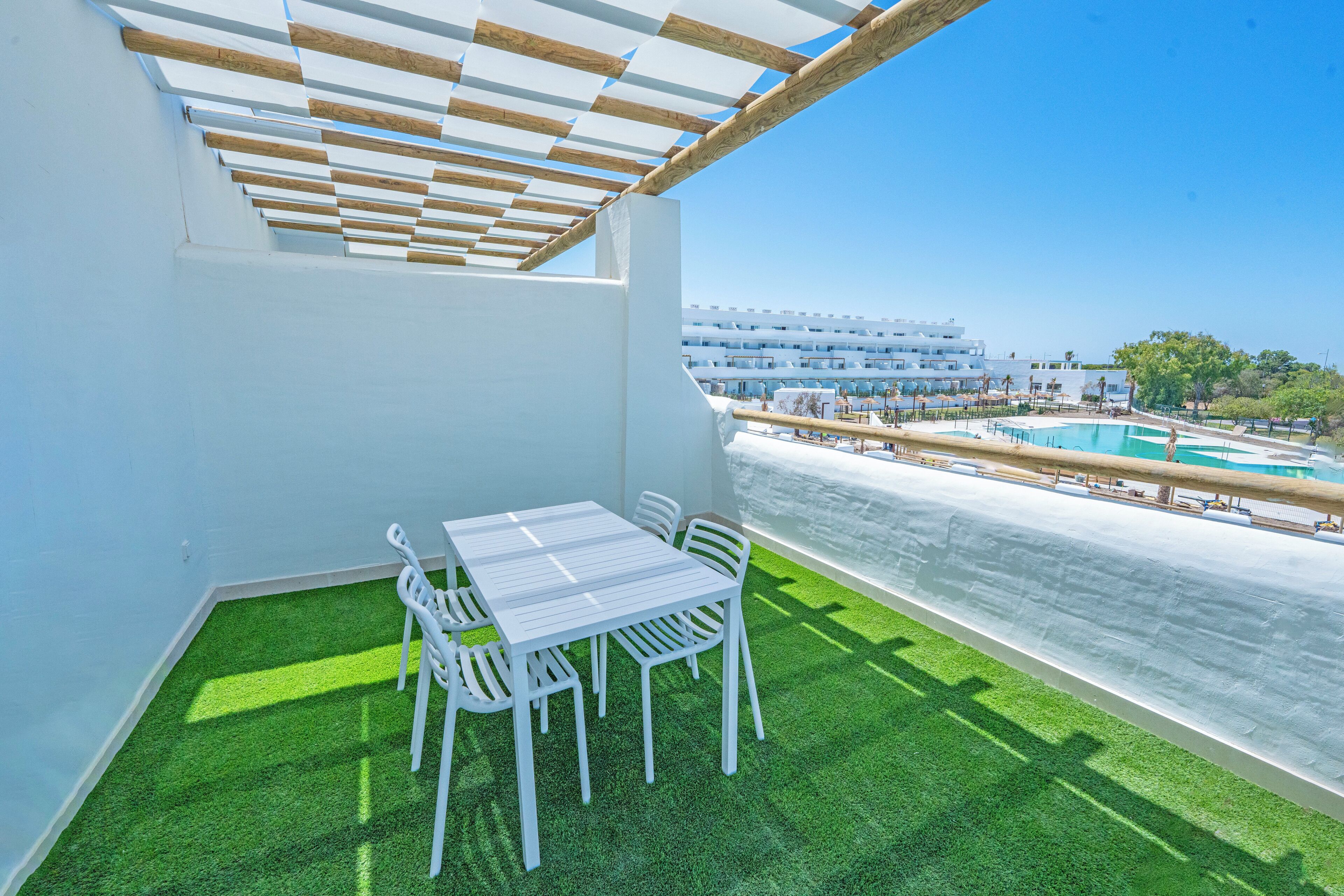 A balcony with a white table and chairs, green artificial grass, a wooden pergola, and a view of a pool and buildings in the background.