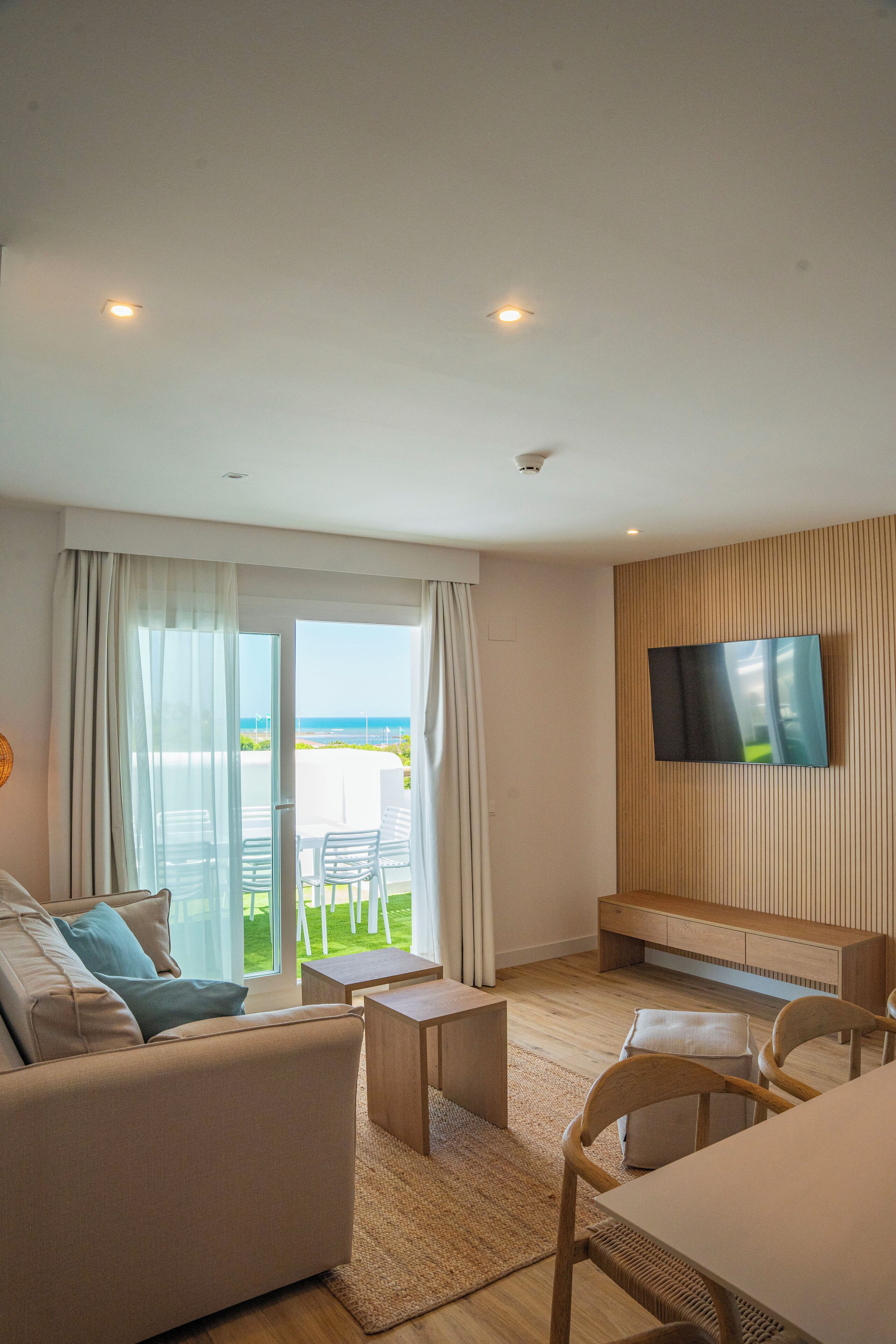 A modern living room with a sofa, a coffee table, and a TV mounted on the wall. There is a large window with curtains, and a view of the outdoors.