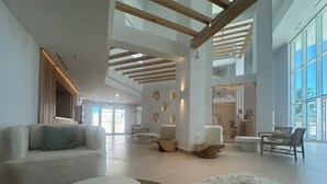 Un vestíbulo espacioso con techo alto, vigas de madera y ventanas amplias. Varias zonas de estar con muebles modernos y un mostrador de recepción.