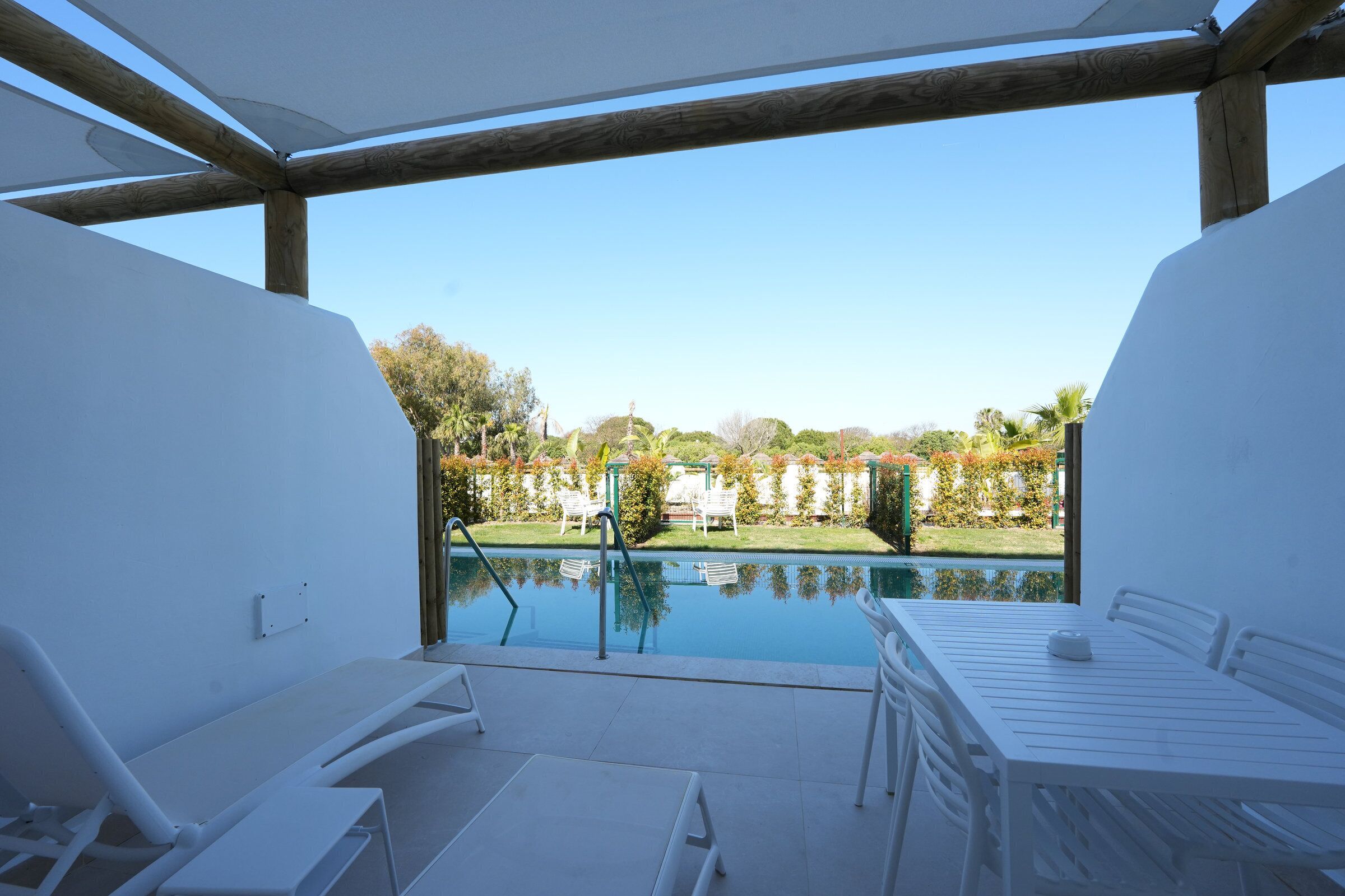A poolside area with a table and chairs, a pool, and a view of a garden and trees.