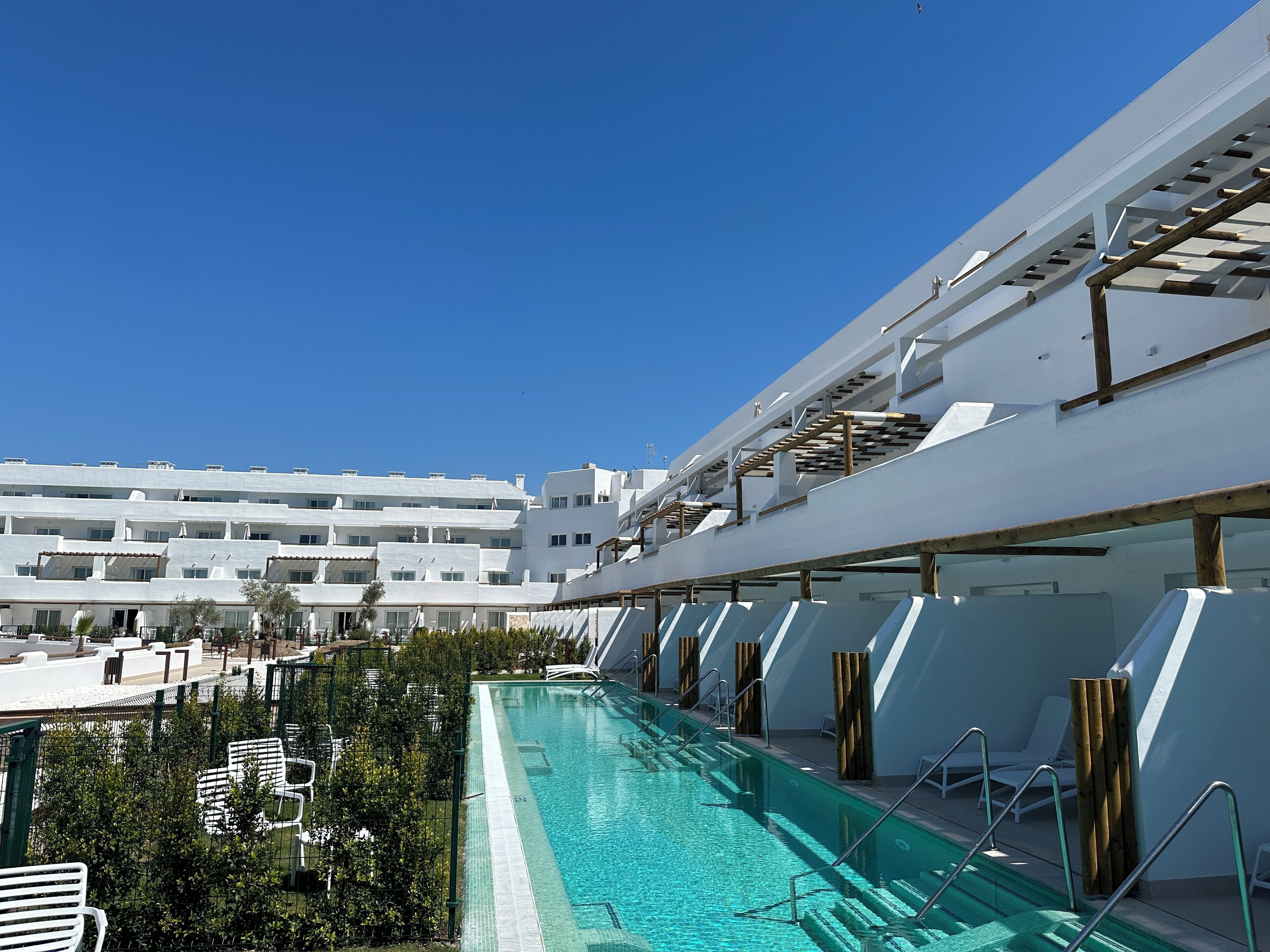 Una piscina con tumbonas y vistas a un edificio blanco con varios balcones.