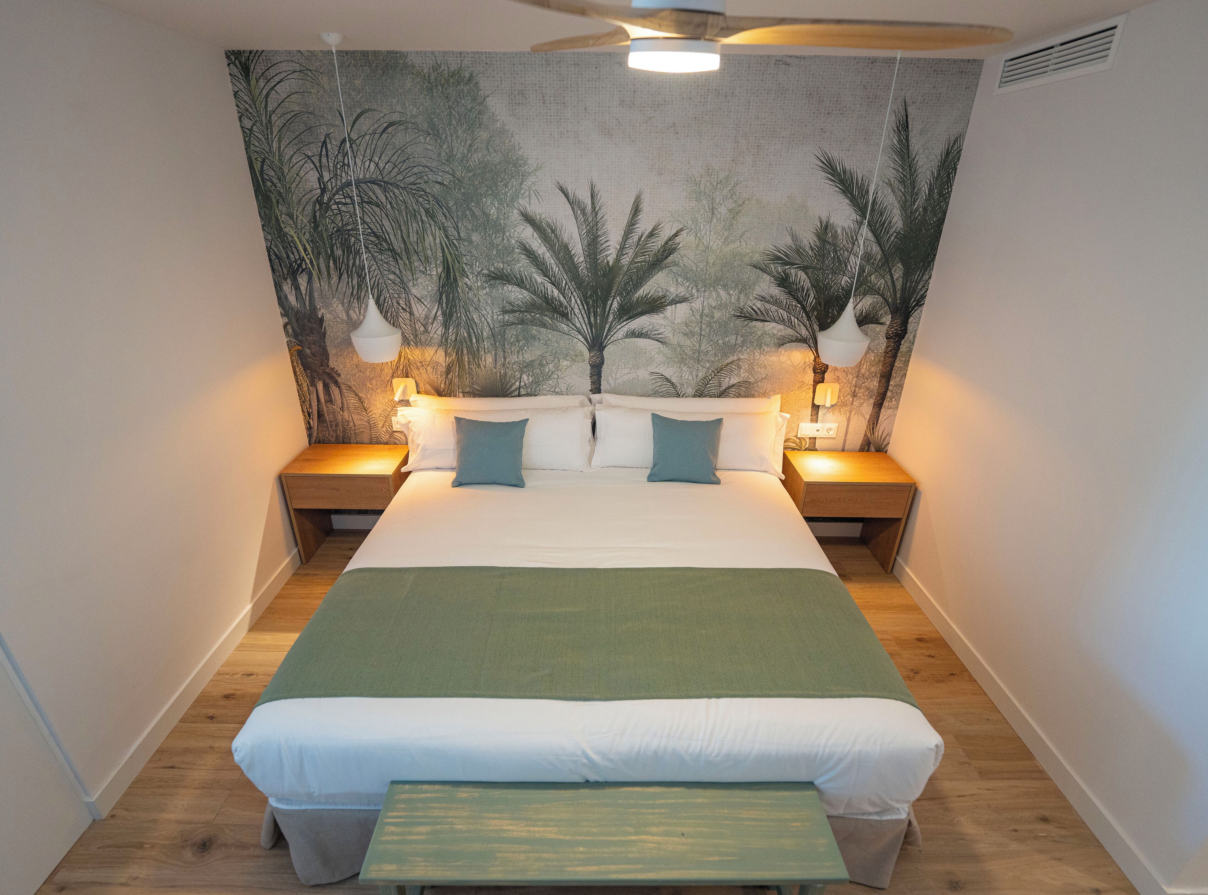 Un dormitorio ordenado con una cama grande, dos mesitas de noche con lámparas, un ventilador de techo y un mural temático tropical.