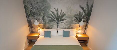 Un dormitorio ordenado con una cama grande, dos mesitas de noche con lámparas, un ventilador de techo y un mural temático tropical.