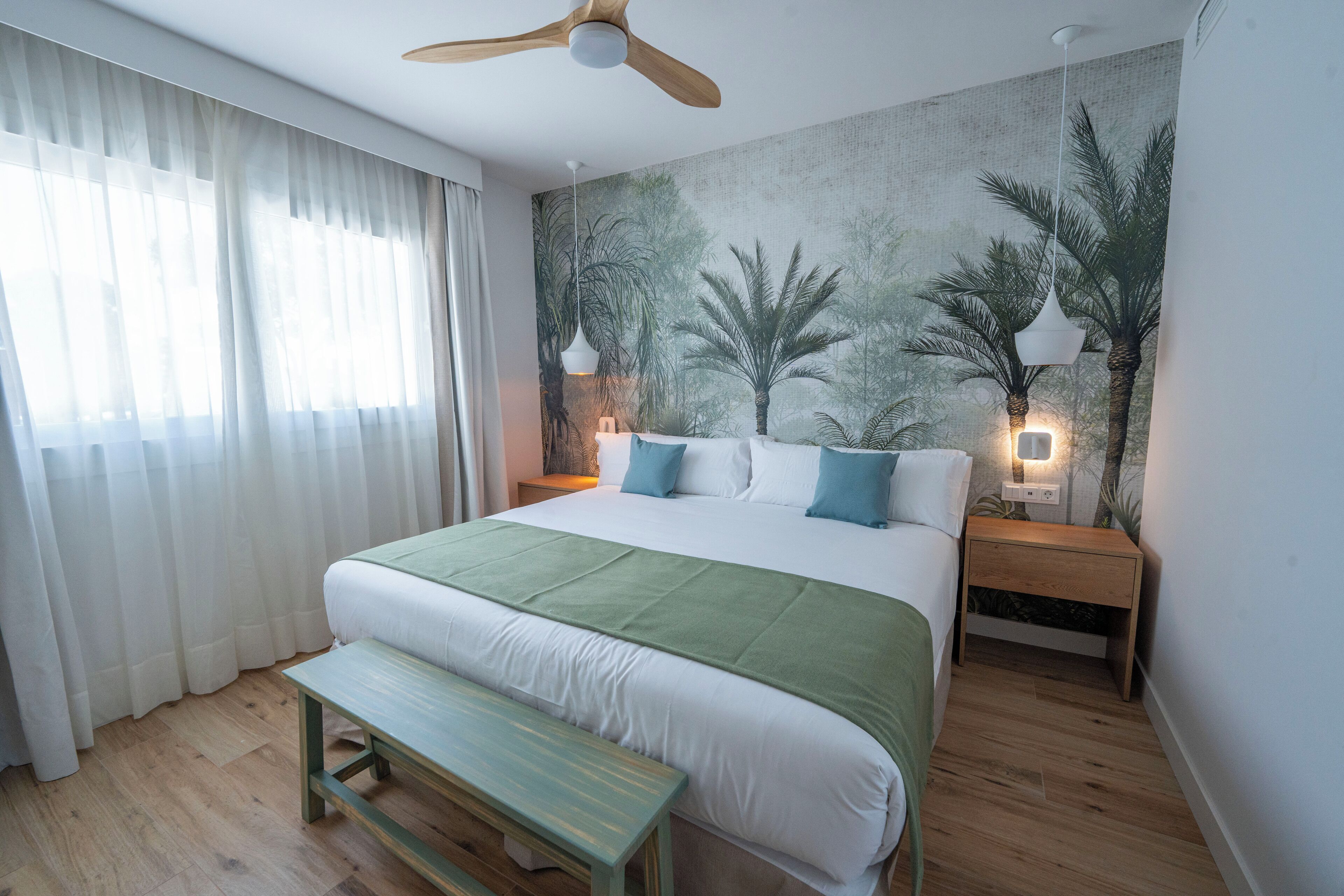 A bedroom with a large bed, a wooden bench, a ceiling fan, and a wall mural of palm trees.