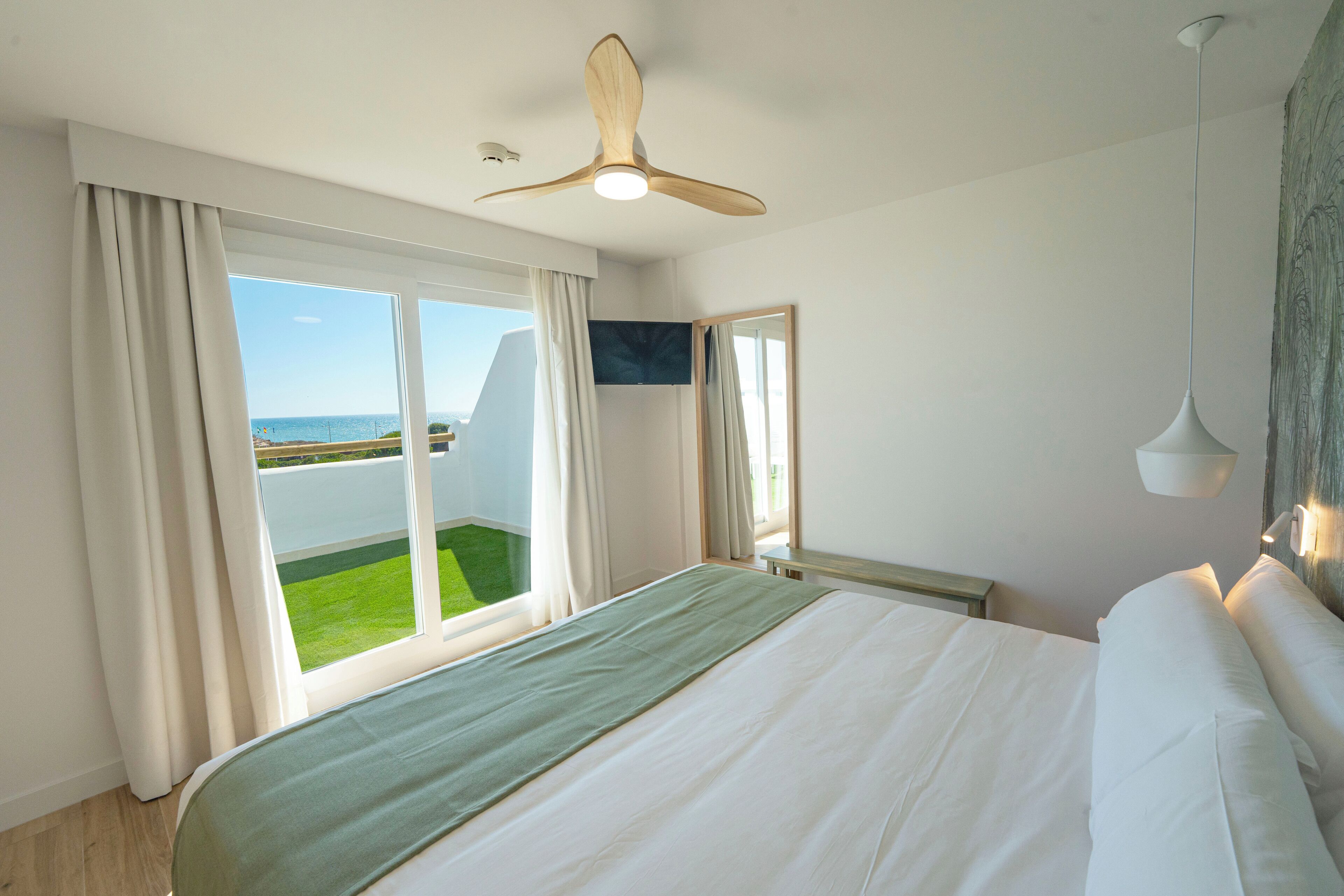A bedroom with a large bed, a ceiling fan, a view of the sea, and a balcony.