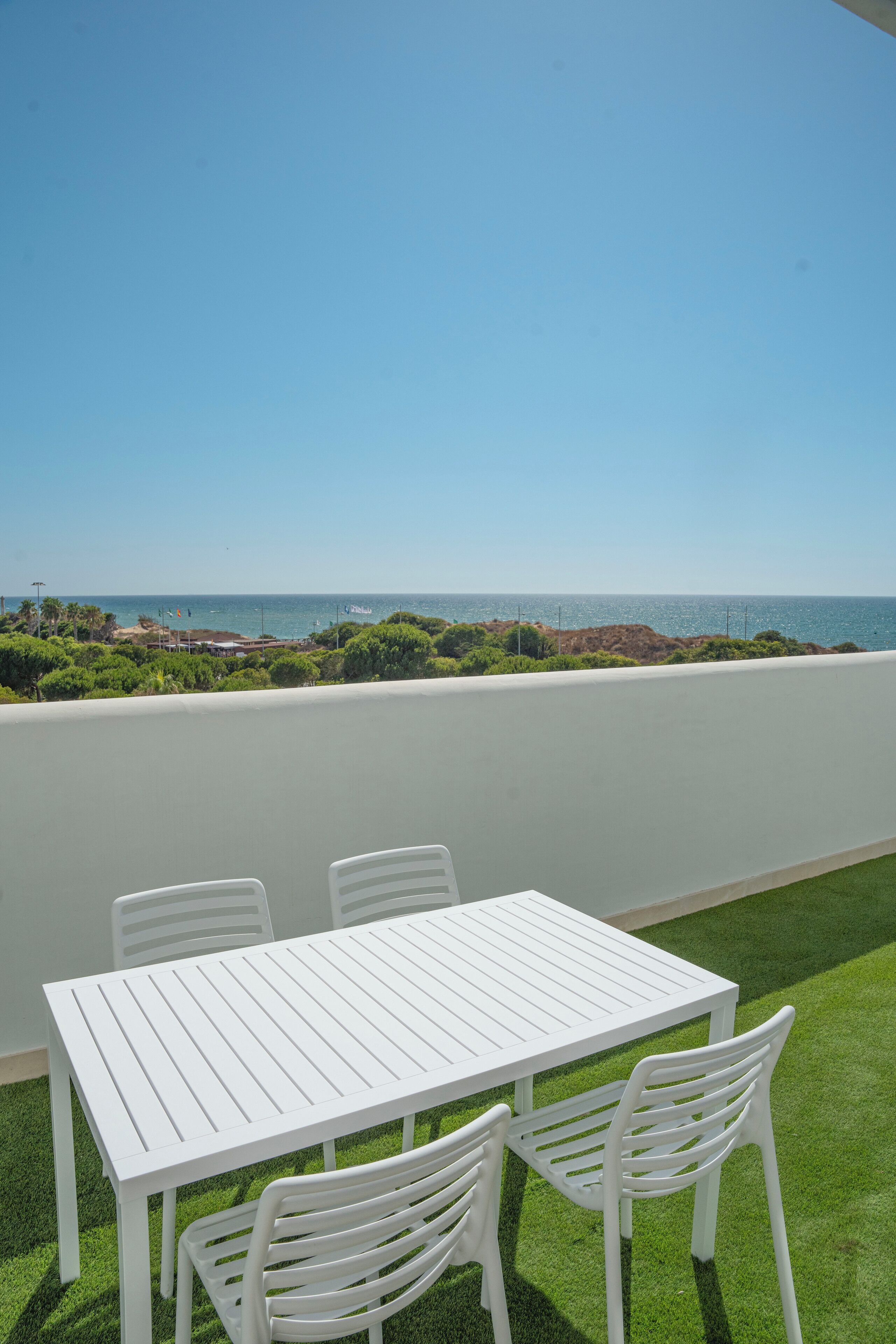 A white table and chairs on a balcony with a view of the sea and greenery.