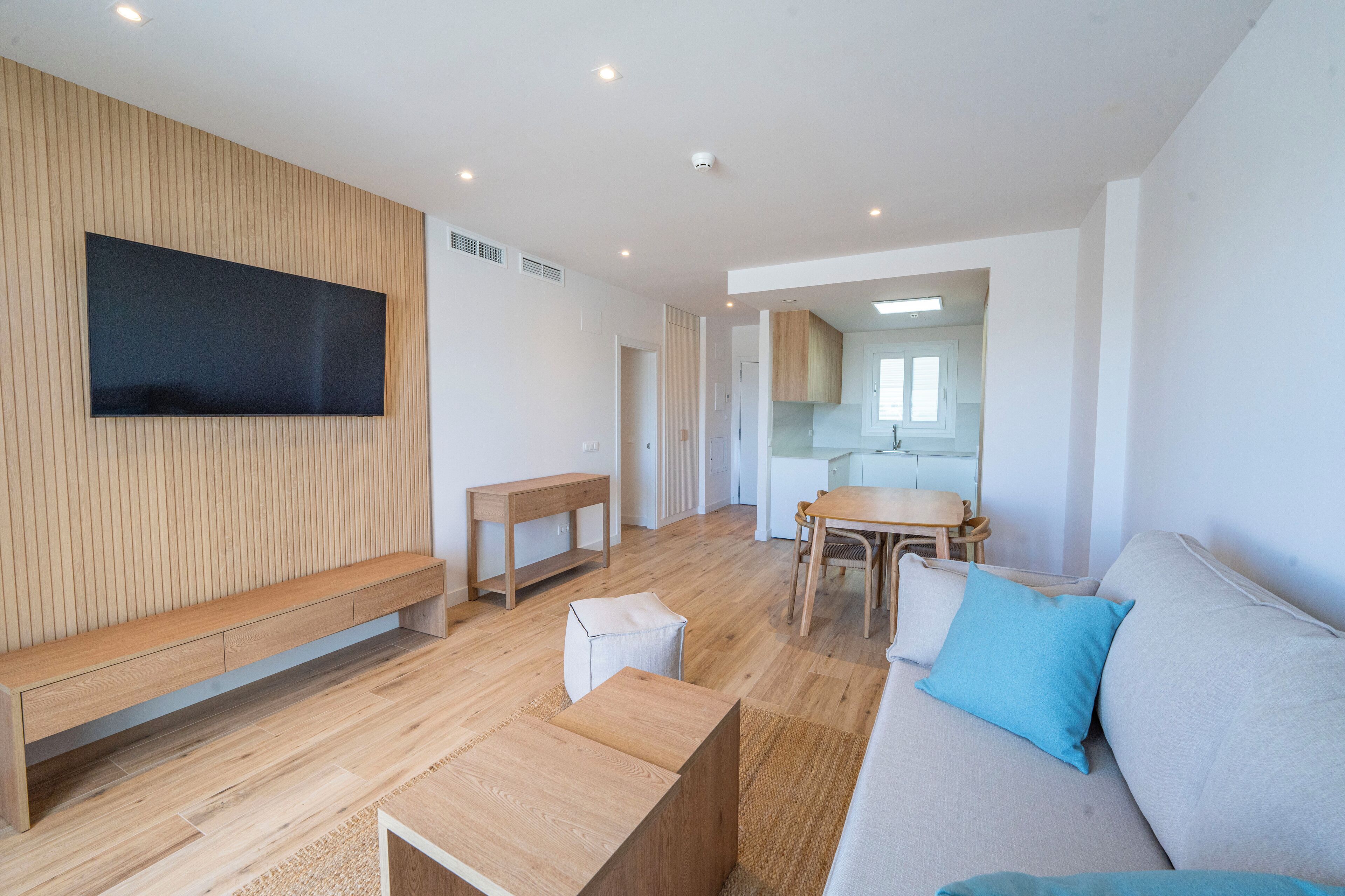 Un salon moderne avec un téléviseur à écran plat fixé à un mur en bois, un banc en bois, un petit pouf, un canapé avec des coussins bleus et un coin repas avec une table et des chaises en bois.