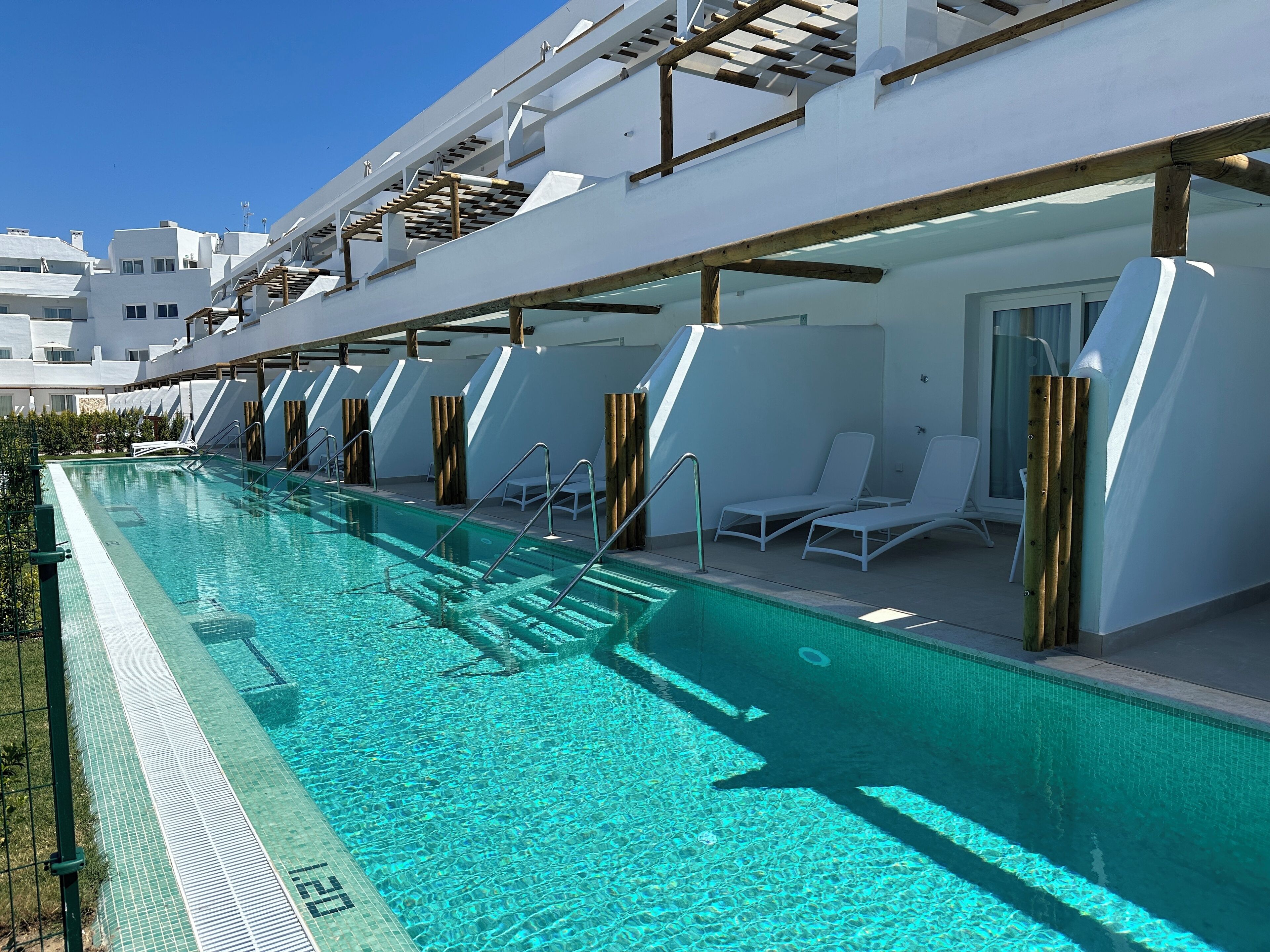 Una piscina con agua cristalina azul, junto a un edificio blanco de diseño moderno, que cuenta con una zona cubierta con sillas de descanso y postes de madera.