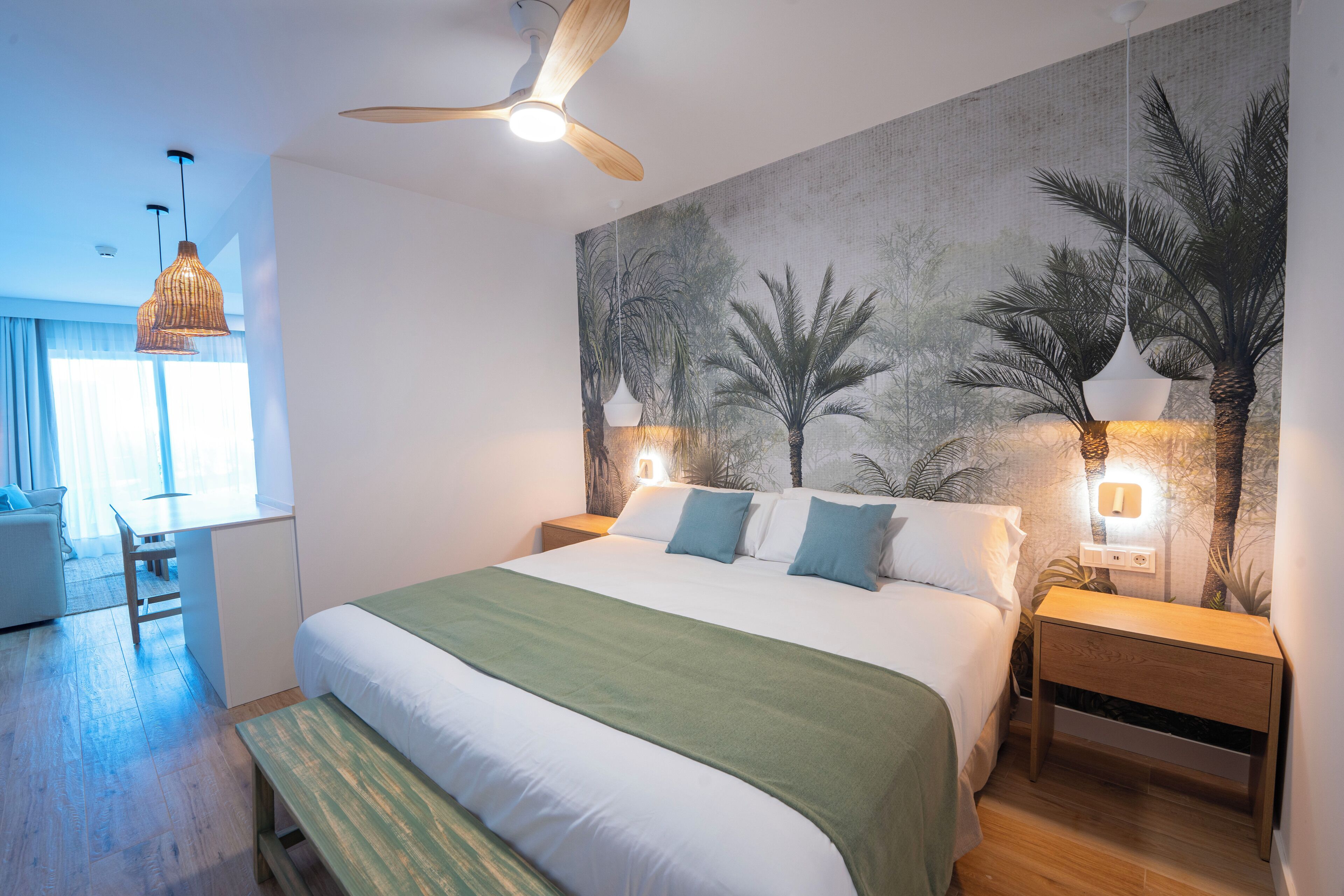 Ein modernes Schlafzimmer mit einem großen Bett, einem Schreibtisch und einem Stuhl. Es gibt einen Deckenventilator, zwei Wandleuchten und ein großes Wandbild von Palmen.
