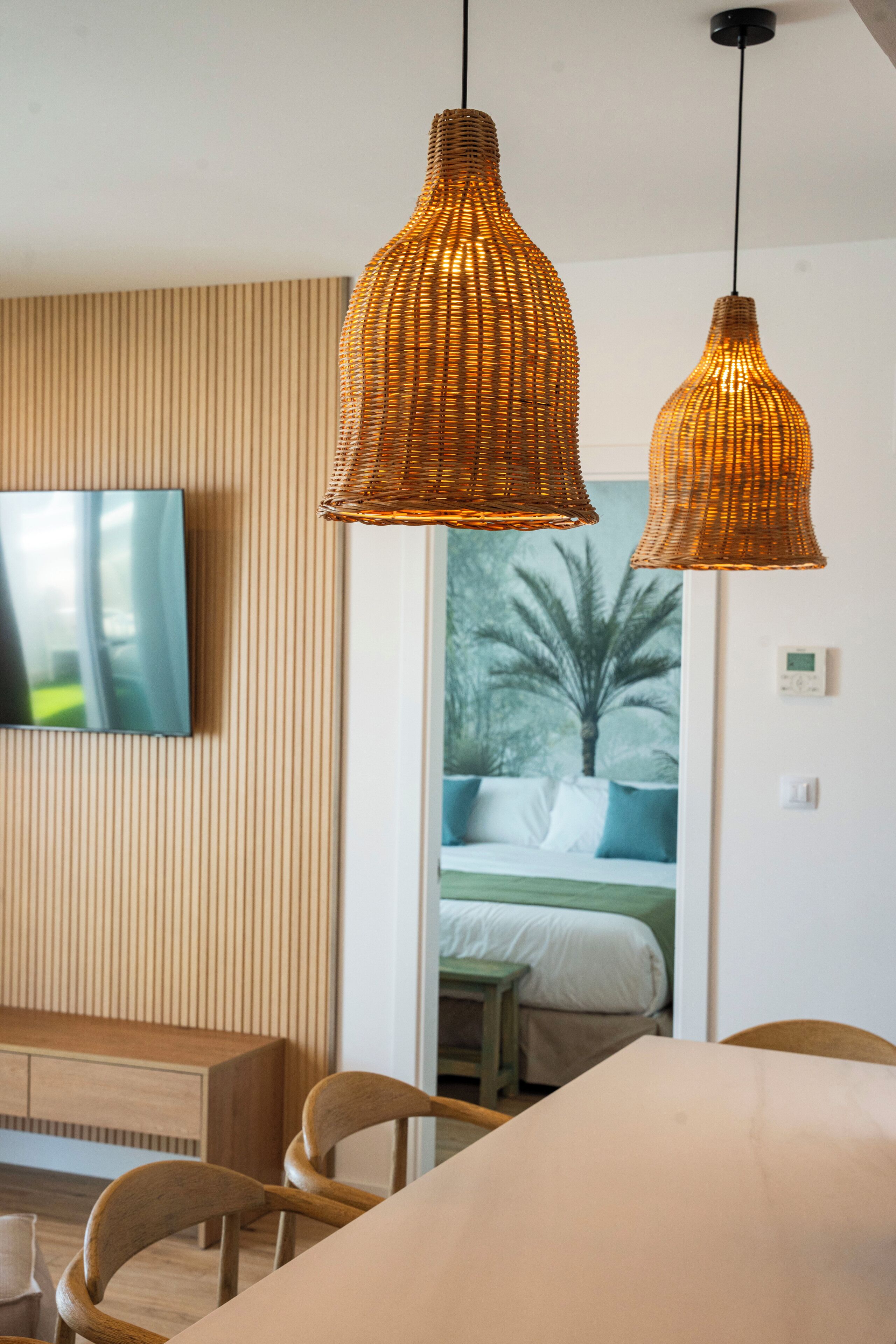 Una habitación moderna con pared de listones de madera, televisor de pantalla plana, lámparas colgantes de mimbre, cama con vista a palmeras y una mesa de comedor de madera con sillas.