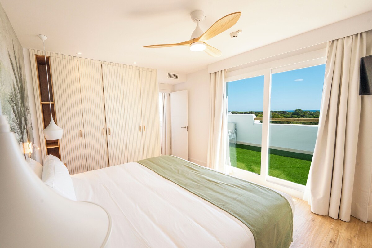 A modern hotel room with a large bed, a ceiling fan, a TV, and a balcony with a view of the greenery and sky.