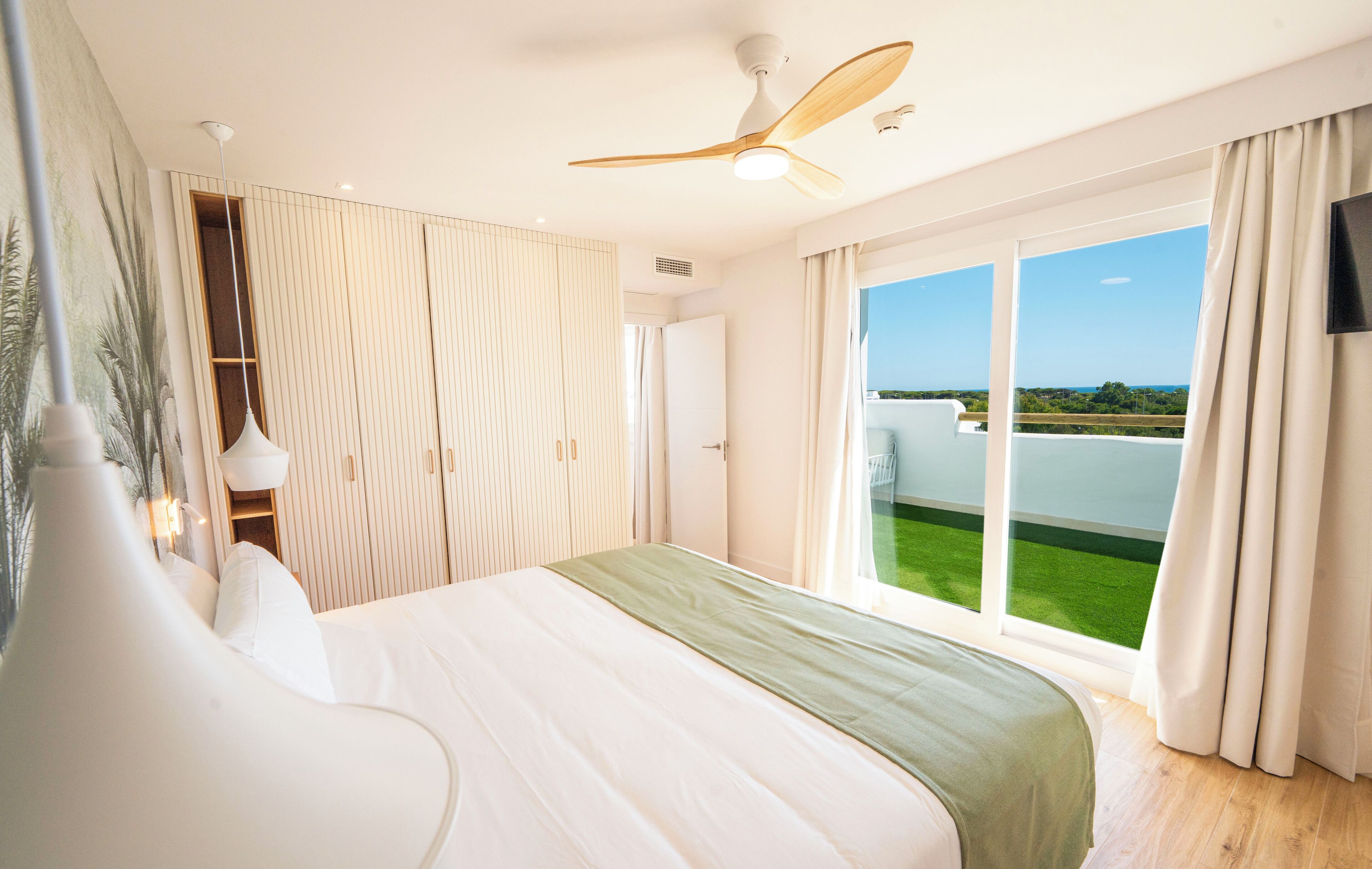Une chambre d’hôtel moderne dotée d’un grand lit, d’un ventilateur de plafond, d’une télévision et d’un balcon offrant une vue sur la verdure et le ciel.