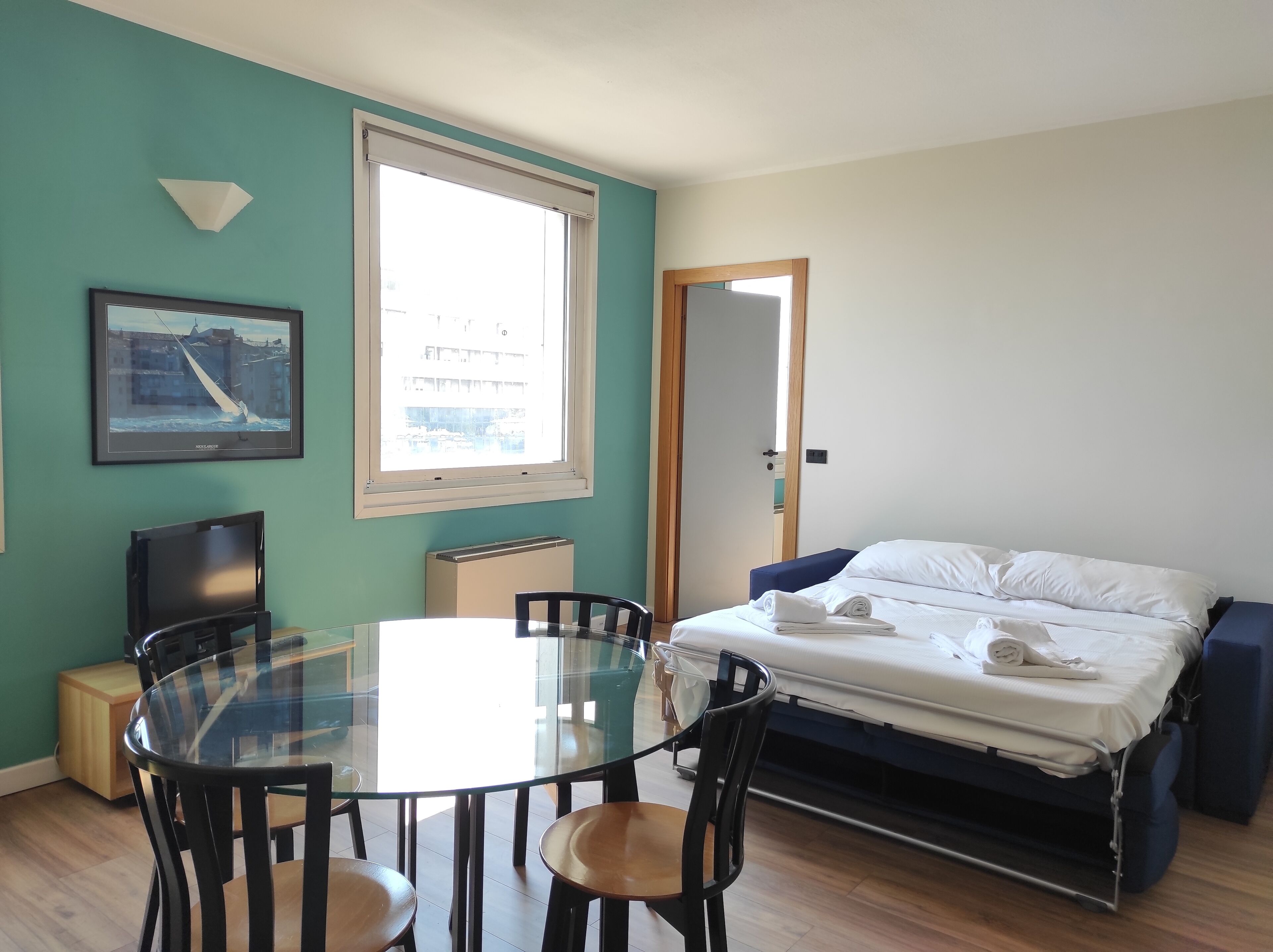 A compact room with a bed, a round glass table with chairs, a framed picture on the wall, and a window with a view of buildings.
