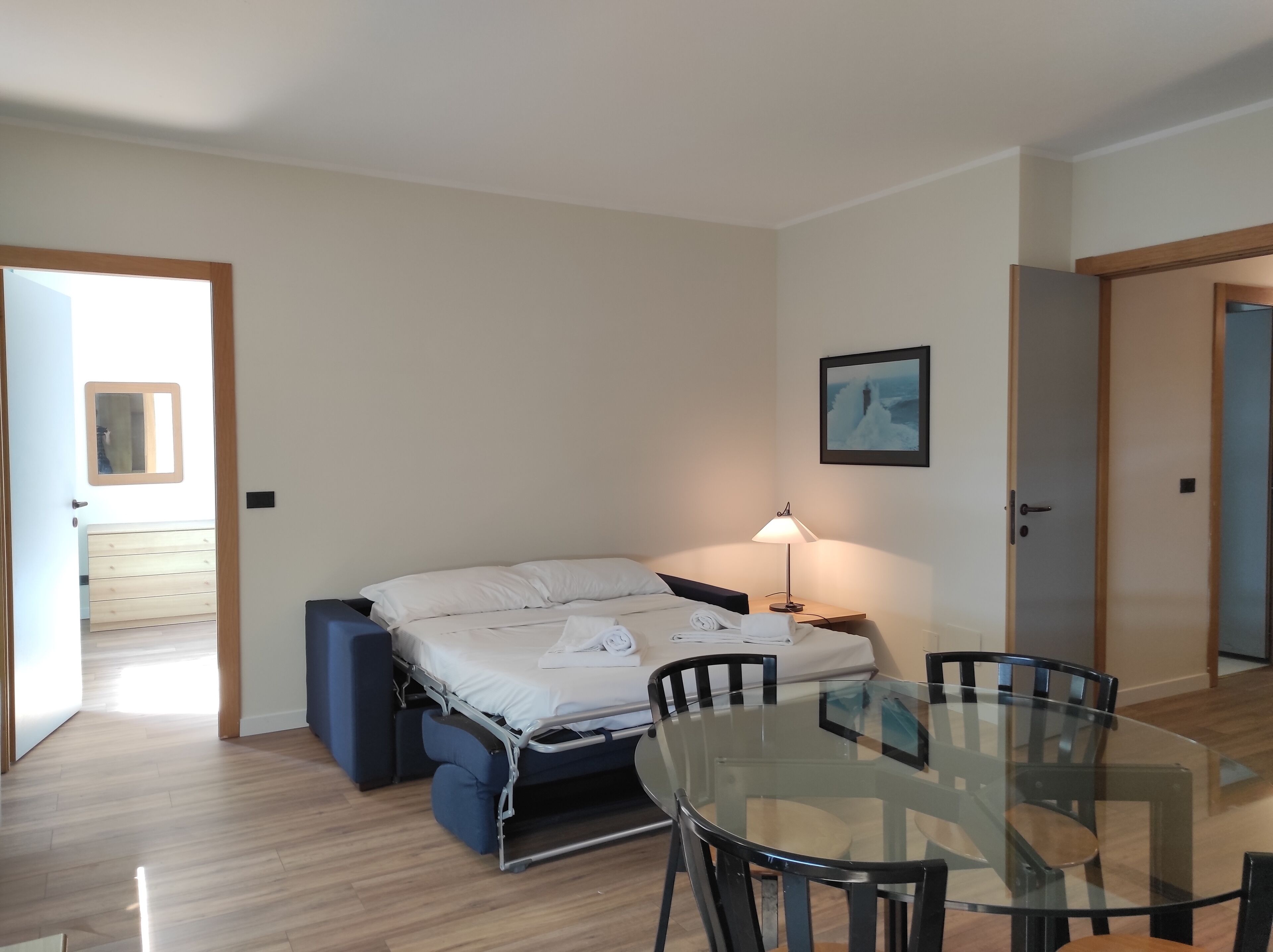A compact room with a bed, a glass table, and chairs, a bedside lamp, and a door leading to another room.