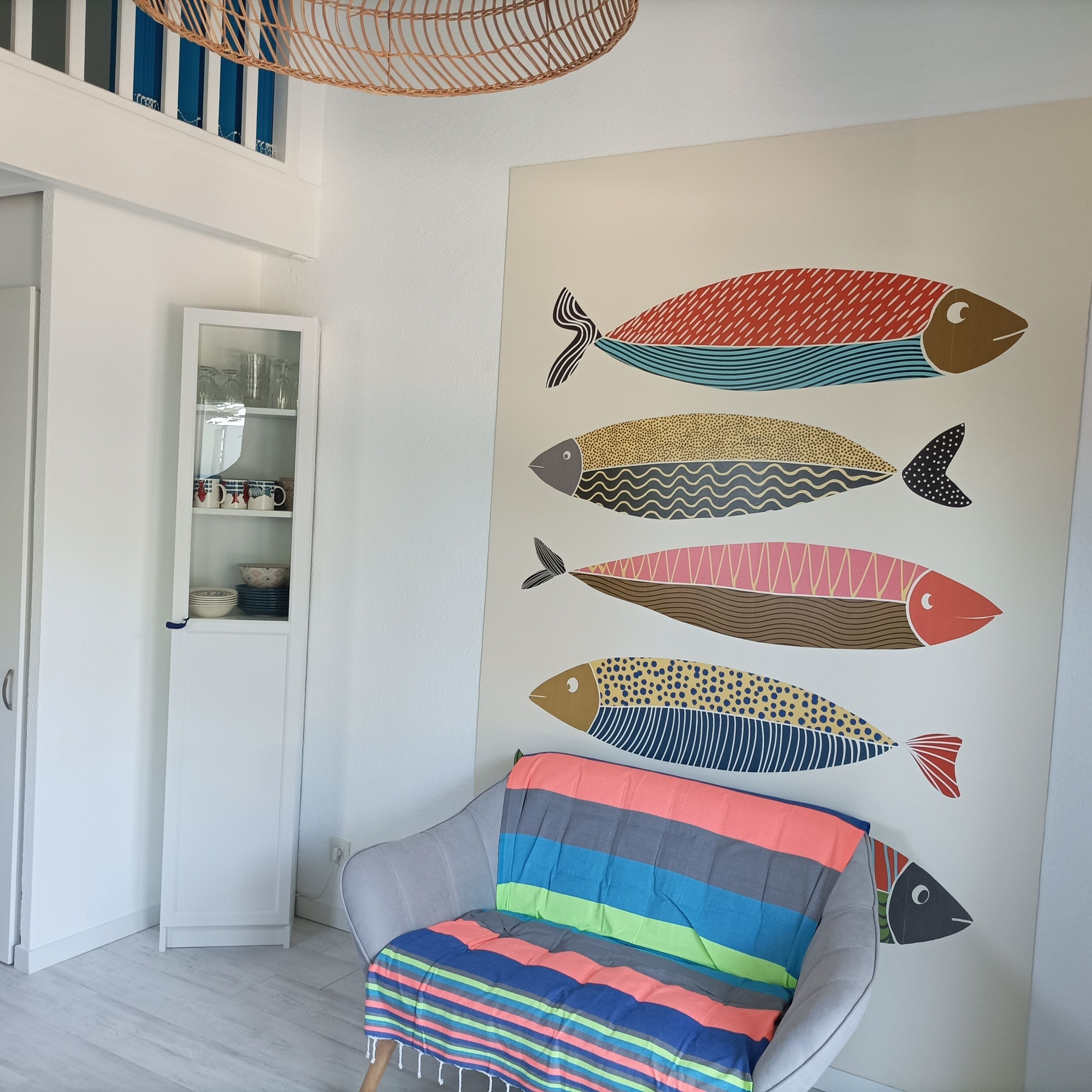 Une pièce avec un mur blanc orné d’une fresque représentant des poissons, un fauteuil gris avec un coussin à rayures et une porte en bois surmontée d’une vitrine.