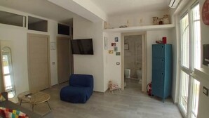 Ein geräumiger Wohnbereich mit einem blauen Sofa, einem kleinen runden Tisch und einem an der Wand befestigten Fernseher. Es gibt einen blauen Schrank und eine Tür zu einem anderen Raum.