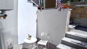 Eine kleine Außenveranda mit einem weißen Tisch und zwei weißen Stühlen, einem Topfpflanzen und einer Treppe, die zu einer oberen Ebene führt.