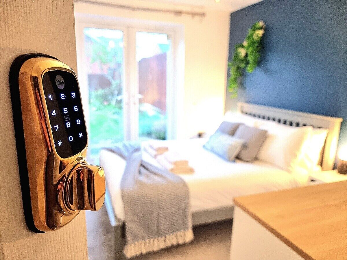Et hotelværelse med en Yale smart lås på døren, en seng med puder, et natbord og et vindue med udsigt til grønne omgivelser.