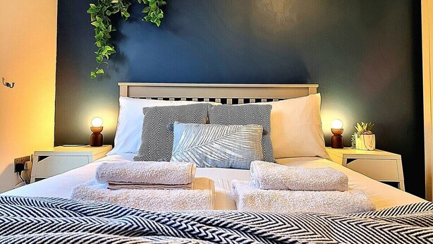 A neatly made bed with multiple pillows and folded towels, a bedside table with a lamp, and a wall-mounted plant. - Spacious House |  Parking | Long Stay Discount (Hamilton)