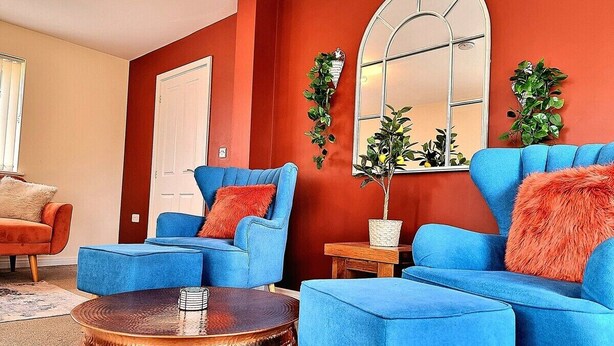 A living room with a red accent wall, blue sofas, a round copper coffee table, and a mirror above a plant. - Detached House w/Free Parking | Long Stay Discount (Leicestershire)
