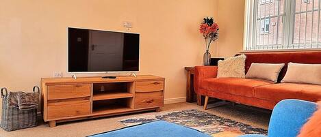 Een woonkamer met een flatscreen televisie op een houten kast, een blauwe bank, een rode bank, en een raam met tralies.